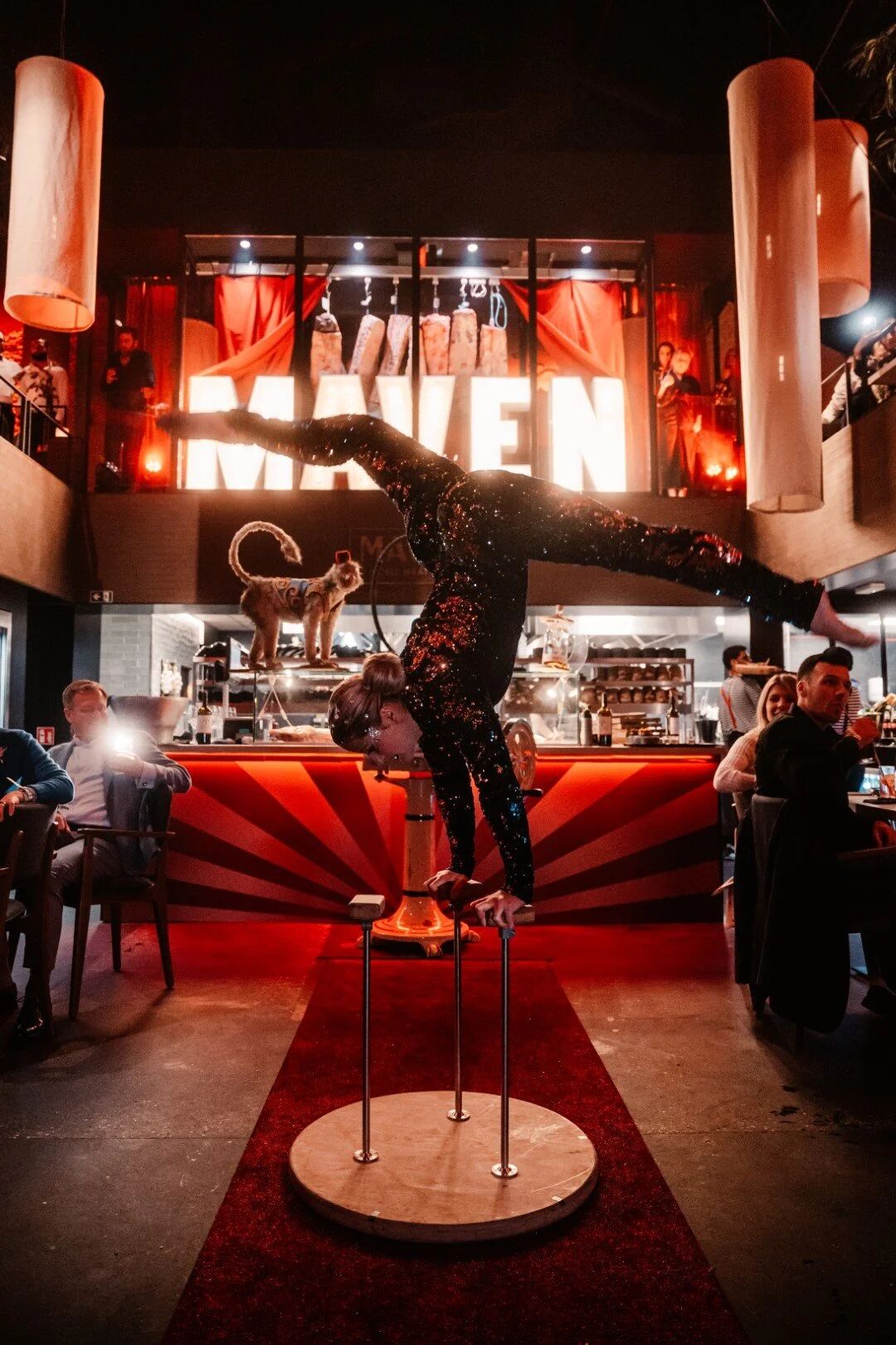 Een acrobate in glitter staat op haar handen voor de bar, onder de lichtletters van Maven.