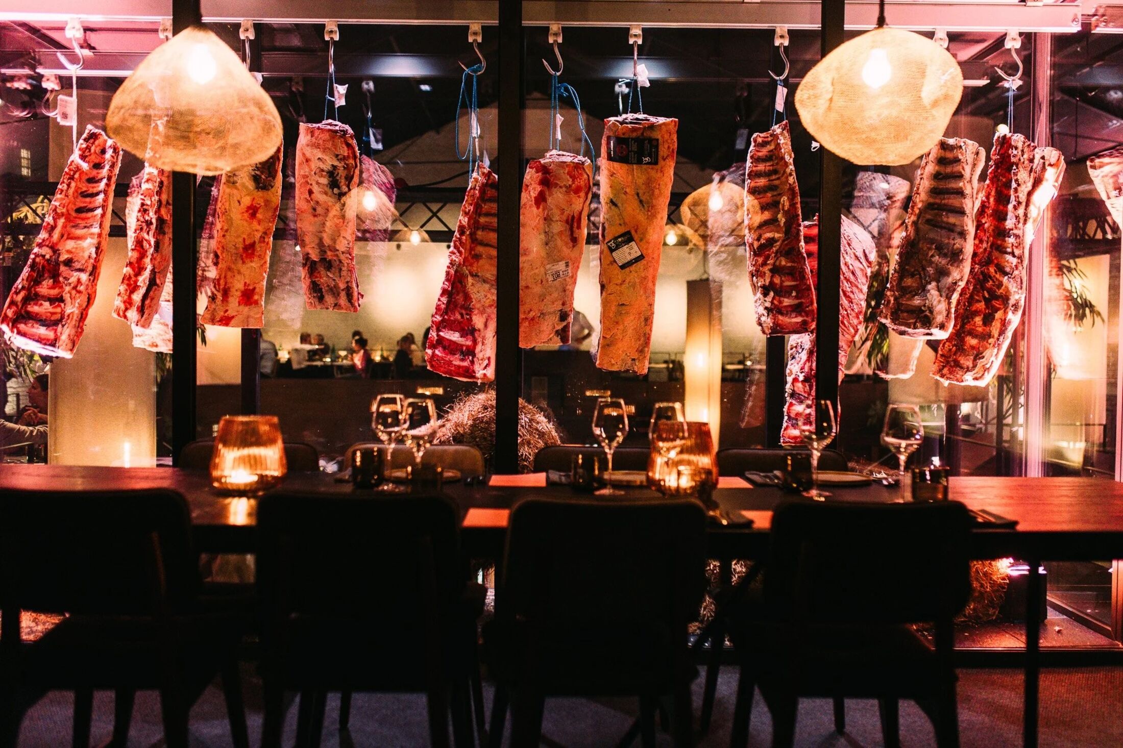 Stukken vlees hangen aan haken boven een gedekte tafel, tussen papieren lantaarns