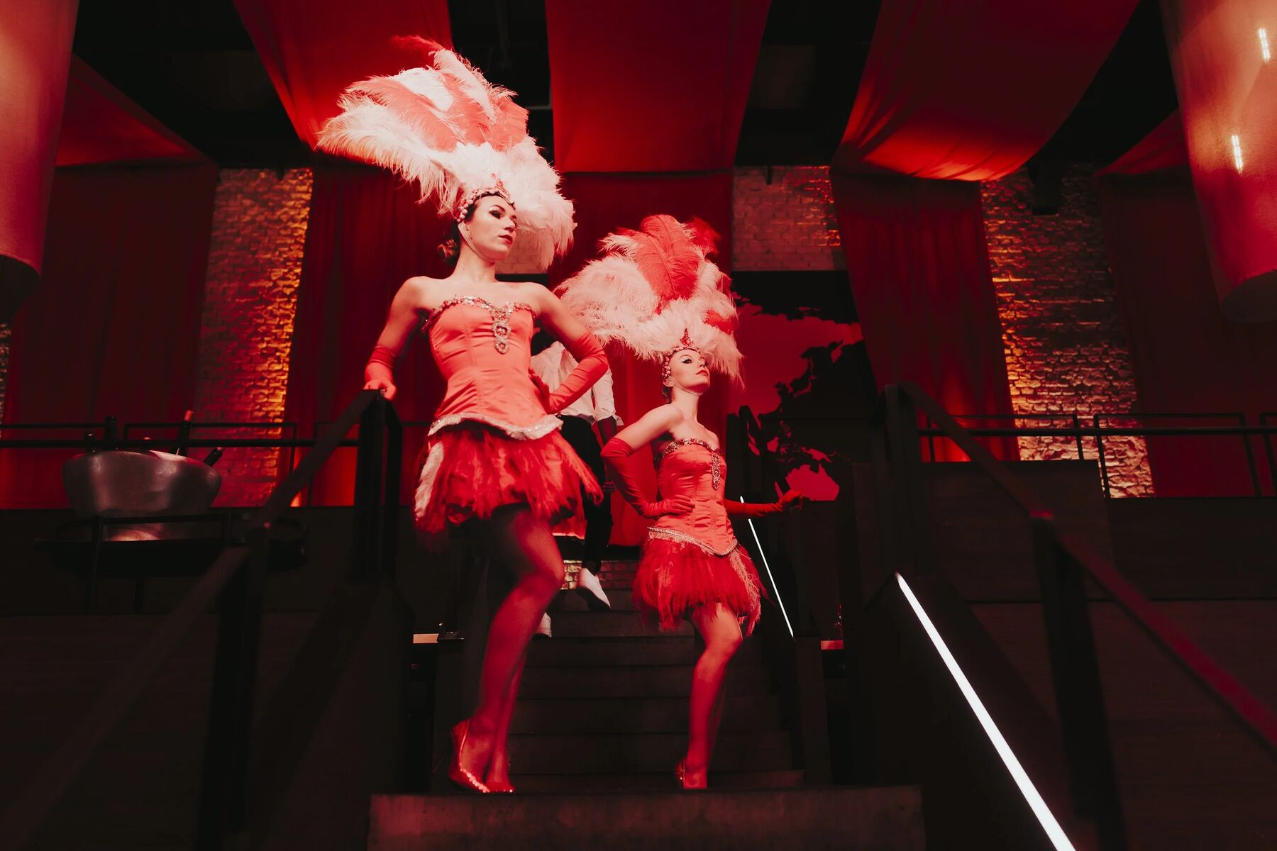 Twee showgirls met pluimen dalen de trap af, in rood licht.